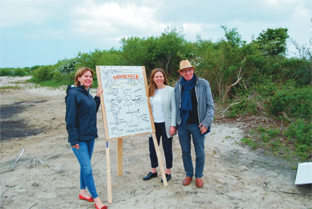 Ron poseert met twee ministers naast een getekende plaat in de duinen.