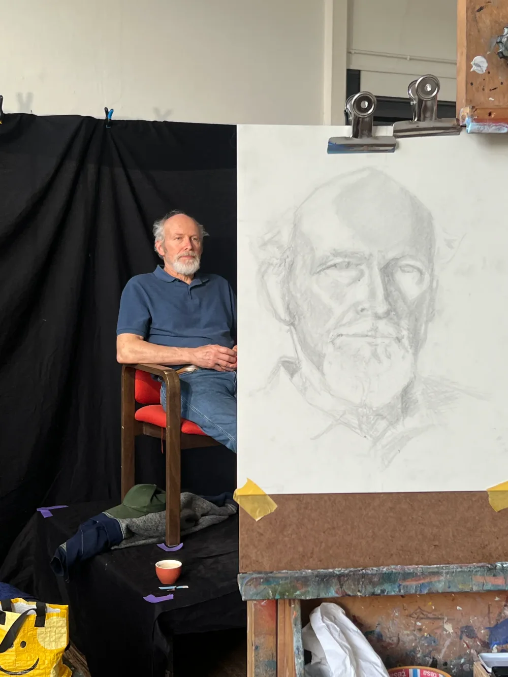 Een oudere man poseert op een stoel; ernaast staat zijn portret in potlood op de ezel.