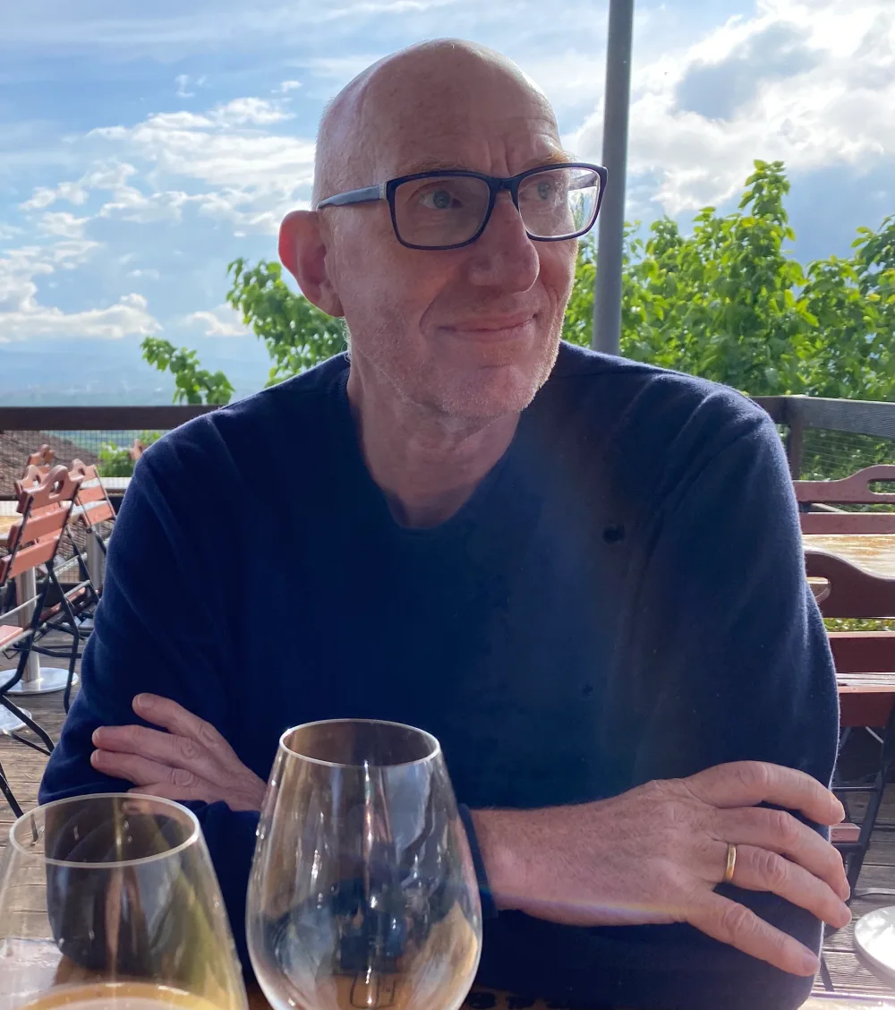Ron Braat zit lachend aan tafel, met een glas wijn voor zich.