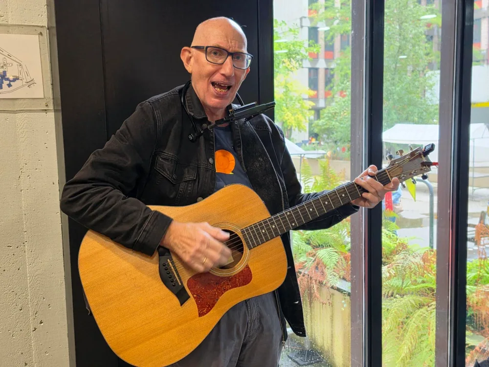 Ron zingt en speelt gitaar met een mondharmonica bij een groot raam.